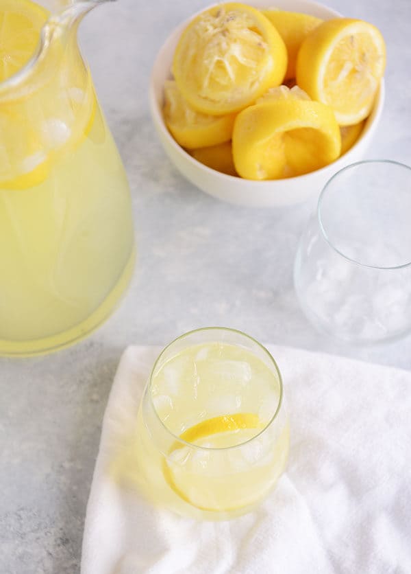 Top view of a glass of lemonade with ice and a bowl of lemons and a pitcher of lemonade beside it.