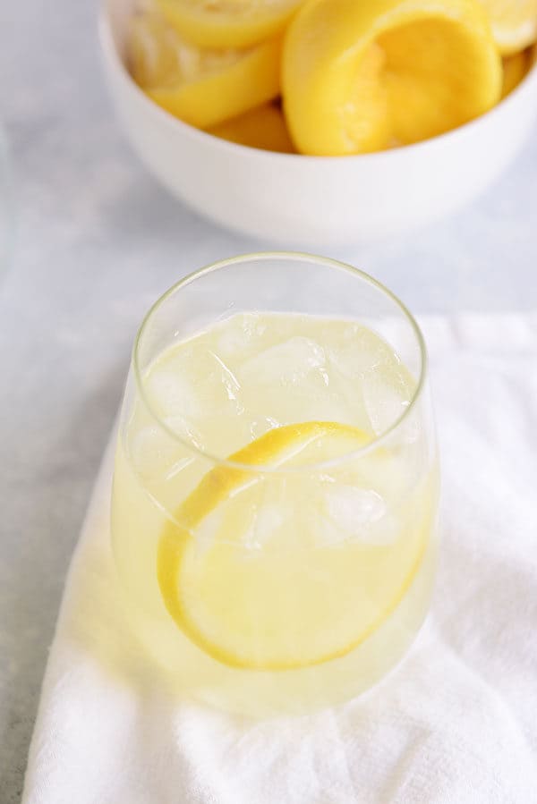 A small glass of lemonade and a bowl of juiced lemons behind it.