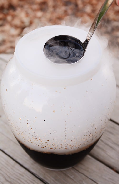 A ladle dipping into a big glass container of homemade root beer with dry ice steam coming out the top.