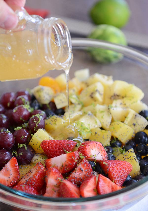 A glass bowl full of chopped fruit with lime zest sprinkled over the top and dressing being poured on the fruit.