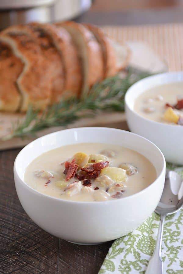 Two bowls of potato soup, topped with crumbled bacon, with fresh sage and a loaf of bread in the background.