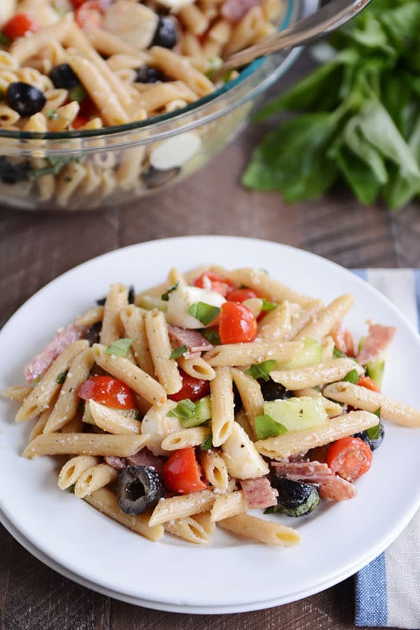 A white plate of penne pasta salad with olives, tomatoes, onions, mozzarella, and basil.