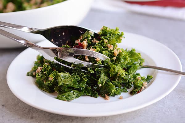 Metal tongs lifting a helping of cranberry kale salad onto a white plate. 