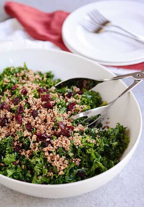 A white bowl full ow cranberry pecan kale salad with metal tongs on the side.