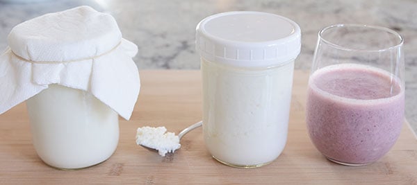 Two mason jars of kefir milk, a spoonful of kefir grains, and a glass of berry kefir milk.