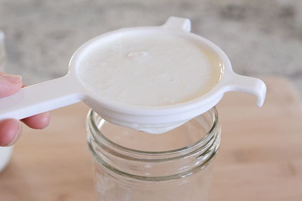 A plastic strainer of kefir grains and milk straining into a mason jar.