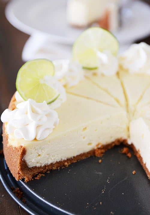 A white cheesecake with a graham cracker crust sliced on a pie plate. Two pieces are missing in the front. Each slice is topped with whipped cream and a slice of lime.