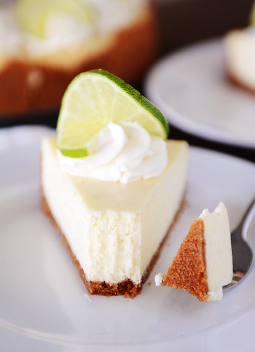 A slice of white cheesecake with one bite taken out on a plate, and the bite on a fork next to it. The cheesecake is topped with whipped cream, and a slice of lime, and has a brown graham cracker crust. 