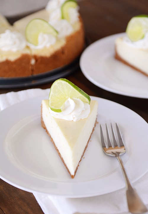 A slice of white cheesecake, topped with whipped cream, and a slice of lime on a white plate with a fork next to it. The other slices of the cheesecake are in the background.