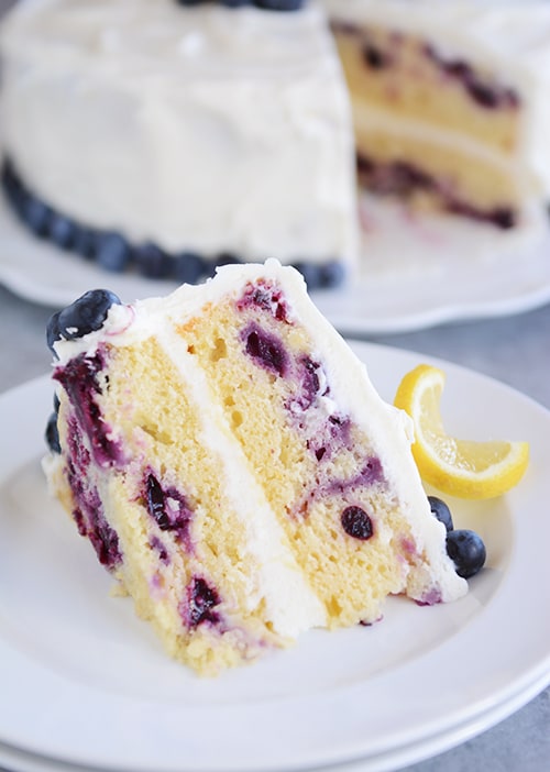 A big slice of lemon blueberry cake on a plate with blueberries and lemon on the side and the rest of the cake in the background.