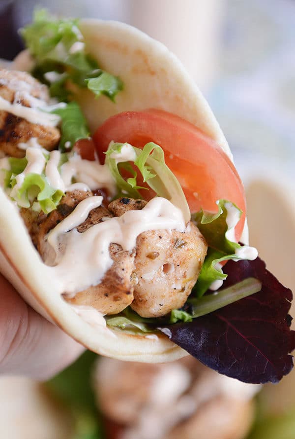 A hand holding a flatbread wrap filled with chicken, lettuce, tomato, and drizzled with sauce.