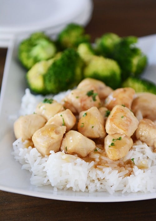 A white platter of cooked rice topped with lemon-glaze chicken next to a helping of broccoli.