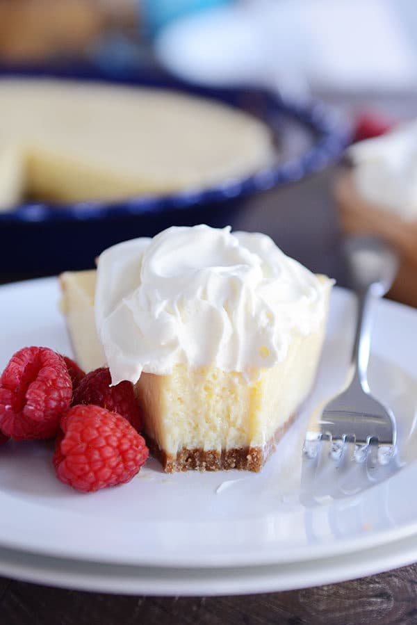 A slice of lemon pie with a bite taken out on a white plate. The pie is topped with whipped cream and there are raspberries on the side. 