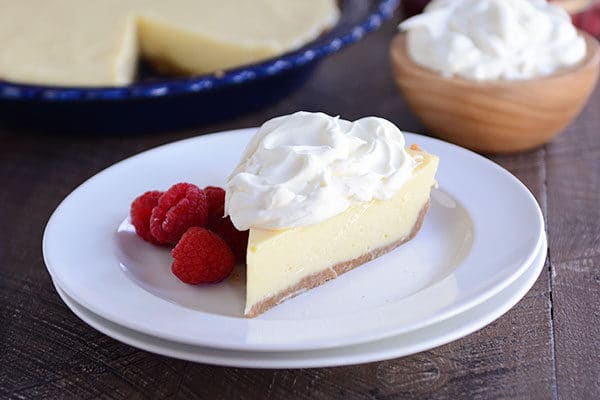 A slice of lemon pie with whipped cream on the top and raspberries on the side on a white plate, with the rest of a pie in a blue pie plate in the background. 