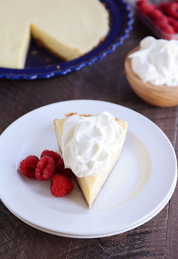 A slice of lemon pie with whipped cream on the top and raspberries on the side on a white plate, with the rest of a pie in a blue pie plate in the background. 