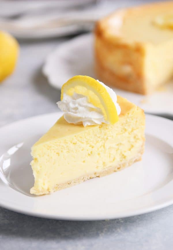 Slice of lemon white chocolate cheesecake on white plate with whipped cream and a slice of lemon on top.