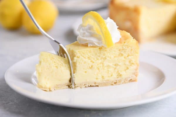 Slice of lemon white chocolate cheesecake on white plate with a fork taking a bite out.