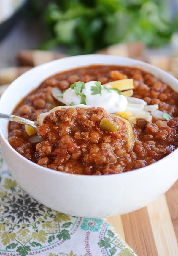 A spoon taking a bite out of a white bowl full of lentil and beef chili. 