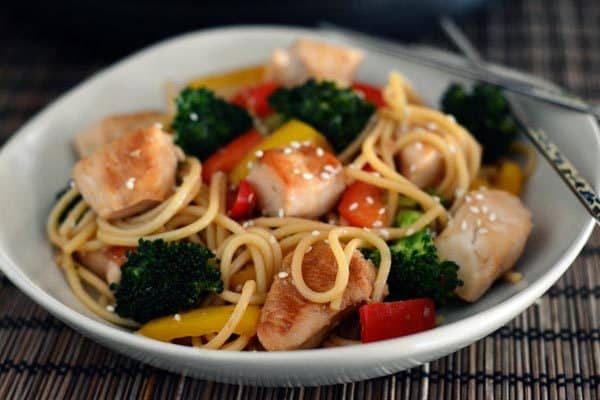 A white bowl of cooked noodles, vegetables, and chicken topped with sesame seeds.