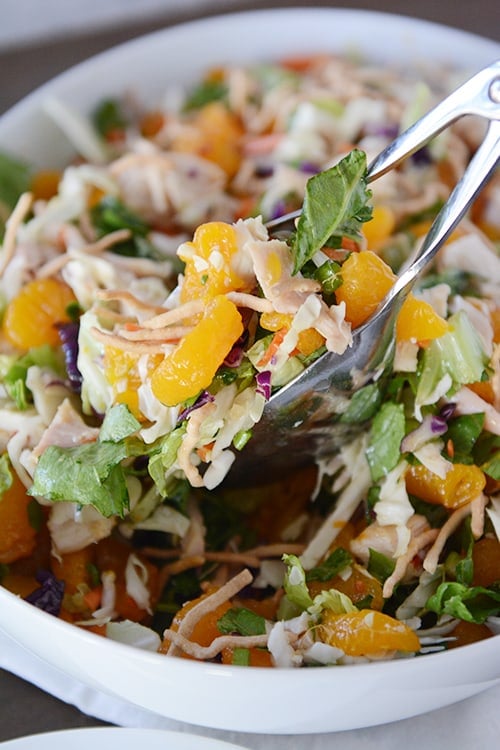 Tongs taking a serving of mandarin orange chicken salad out of a white bowl.