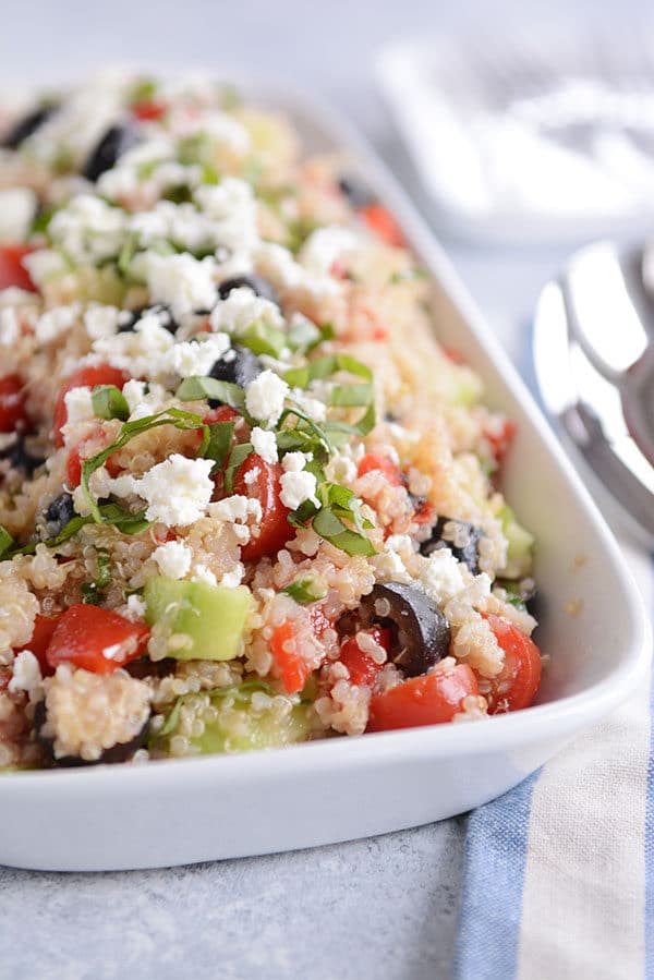 A white dish with cooked quinoa, tomatoes, olives, cucumbers, feta, and basil.