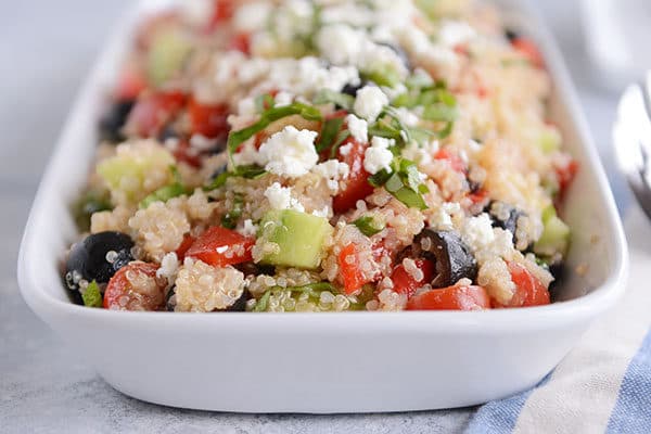 A white dish full of cooked quinoa with chopped cucumbers, olives, and tomatoes and crumbled feta on top.
