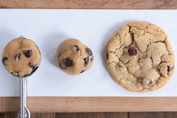 A cookie scoop full of raw cookie next to another ball of raw dough and a baked cookie.
