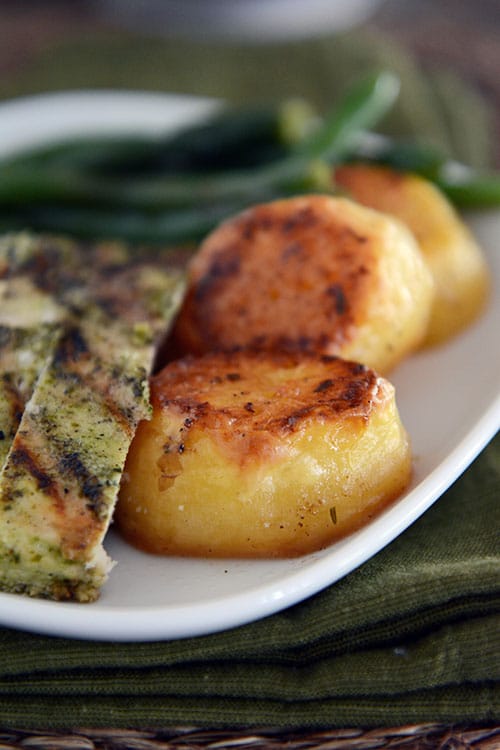 A white plate with golden brown cooked potatoes, slices of pesto chicken, and cooked green beans. 