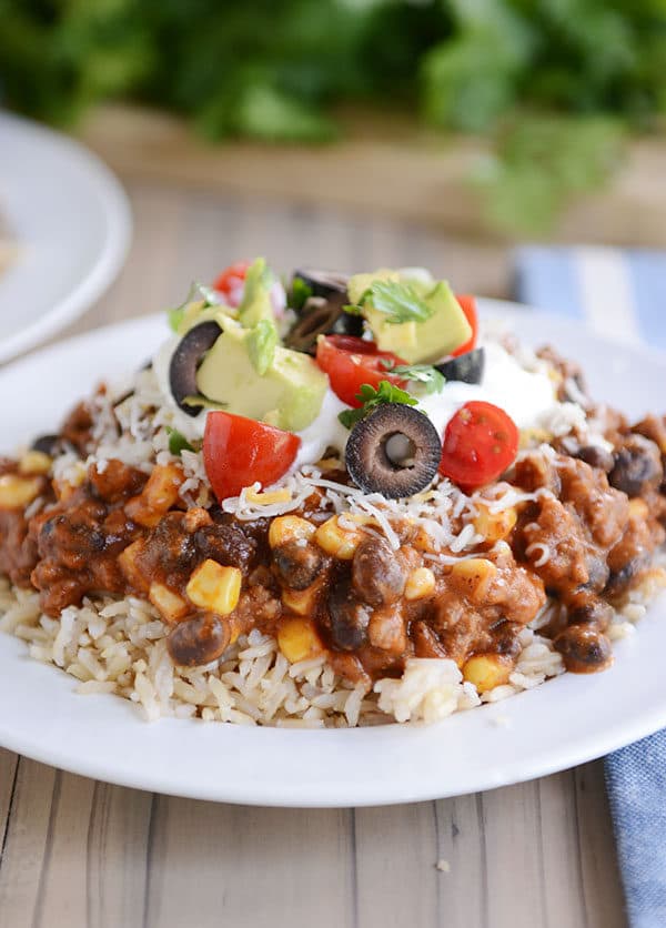 A white plate of cooked rice topped with a Mexican beef and corn mixture, tomato, olive, avocado, and sour cream.