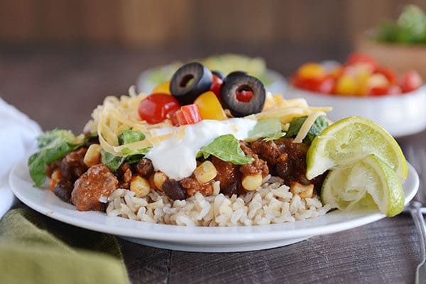 A white plate of cooked rice topped with a beef Mexican haystack mixture topped with taco toppings.