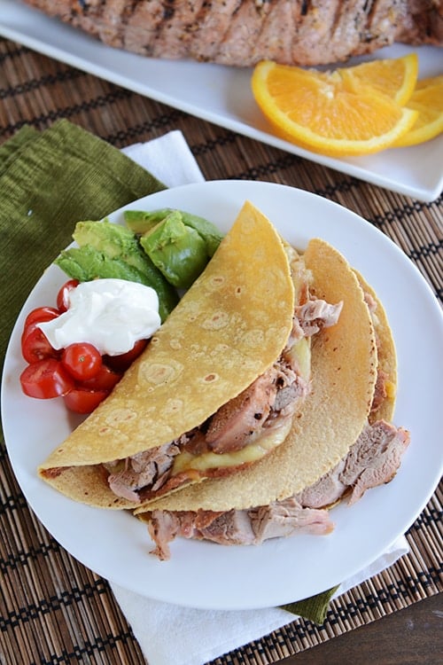 Two pork tacos on a white plate with cherry tomatoes, sour cream, and diced avocado.
