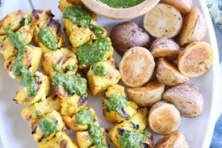 Top down view of white platter with Moroccan chicken skewers, green sauce and roasted potatoes.