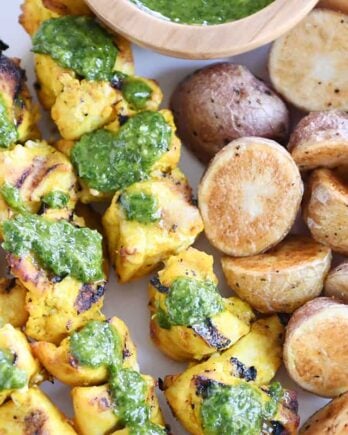 Top down view of white platter with Moroccan chicken skewers, green sauce and roasted potatoes.