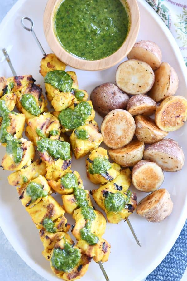 Top down view of white platter with Moroccan chicken skewers, green sauce and roasted potatoes.