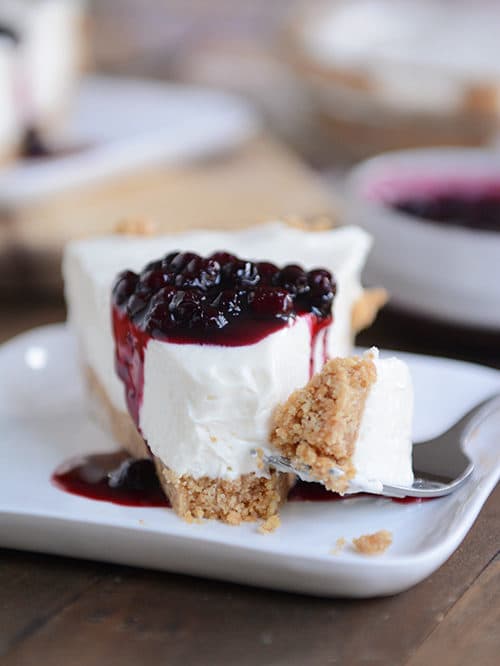 A slice of graham cracker crust no-bake cheesecake, with one bite on a fork on the side ready to eat, and blueberry sauce drizzled on top of the slice. 