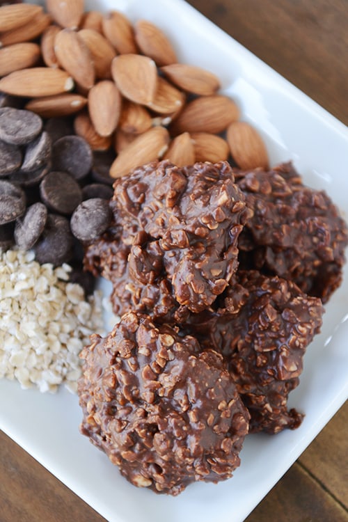 A white tray of no-bake chocolate oat cookies next to piles of chocolate chips, almonds, and oats.