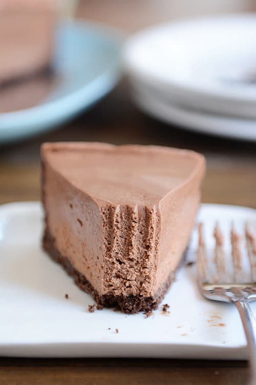 A slice of chocolate cheesecake with chocolate crust with one bite taken out, on a white plate.