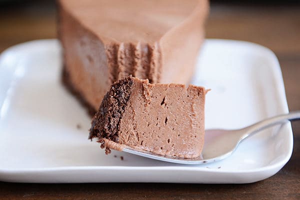 A fork taking a bite out of a chocolate cheesecake with a chocolate crust on a white plate. 