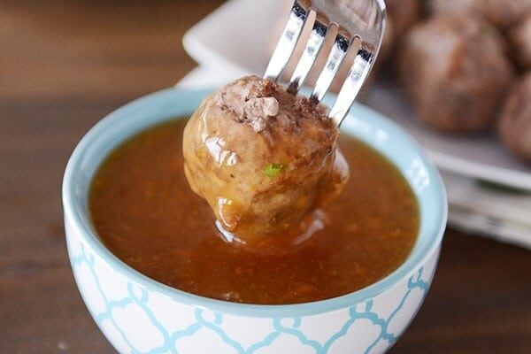 A cooked meatball getting dipped into a bowl of orange sauce. 