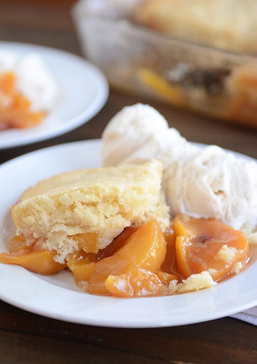 Peach cobbler with a biscuit topping, and two scoops of vanilla ice cream on a white plate.