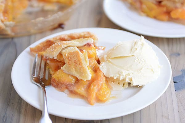 A slice of peach pie and fork next to a scoop of vanilla ice cream on a white plate.