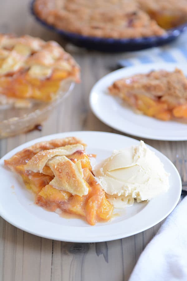 A white plate with a slice of peach pie and ice cream in front of two pie dishes of pie.