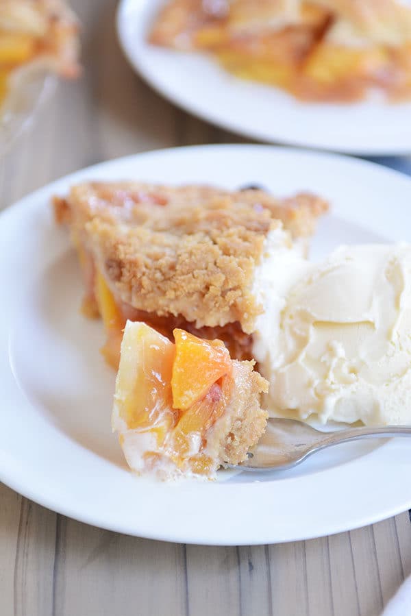 A fork taking a bite out of a slice of fresh peach pie next to a scoop of vanilla ice cream on a white plate.