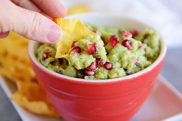 Ted bowl full of guacamole with pomegranate seeds in it and a person taking a bite out with a chip.