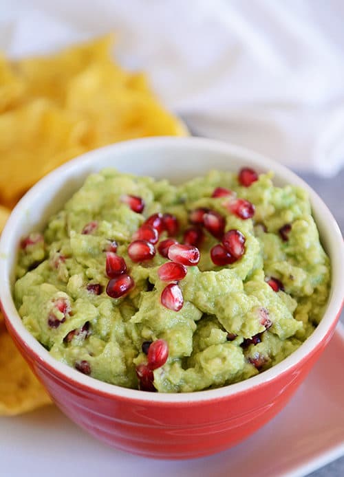 Bowl of pomegranate seed studded guacamole with tortilla chips in the background.