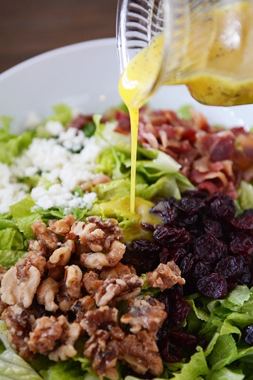 Vinaigrette being poured over a romaine salad with nuts, craisins, feta, and bacon.