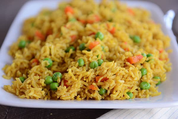 A white platter full of yellow-tinted rice with peas and carrots throughout. 
