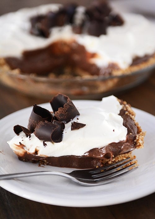 A slice of chocolate pudding pie on a white plate in front of the rest of the pie.