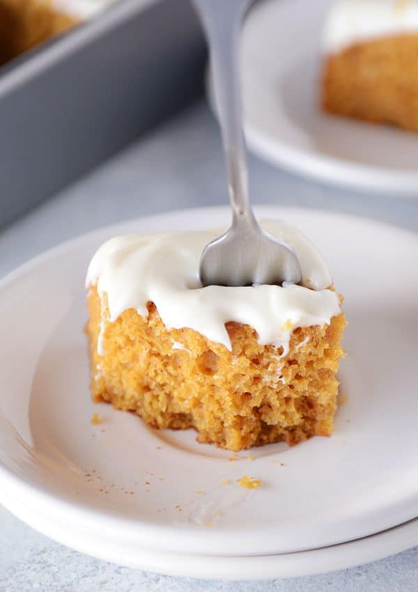 A piece of pumpkin cake with frosting, a fork stuck in the middle, and a bite taken out on a white plate.