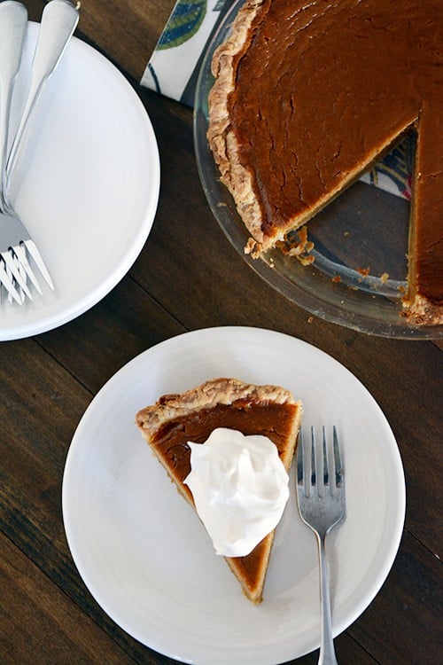 Top view of a pumpkin pie with a missing slice, and the slice taken out on a white plate with a dollop of whipped cream.